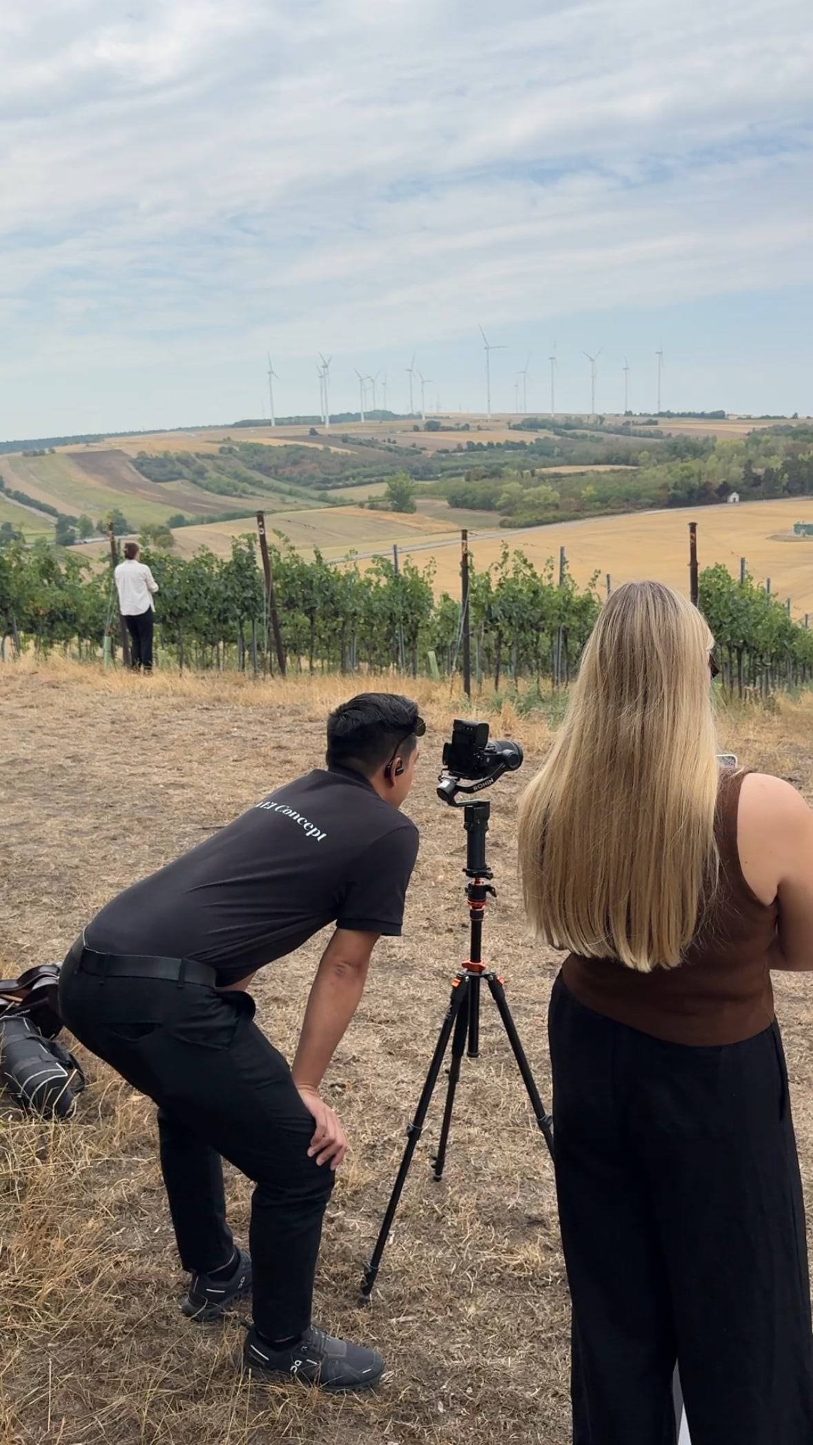 Videograf mit Stativ im Weingarten, daneben die Winzerin, im Hintergrund Weinberge